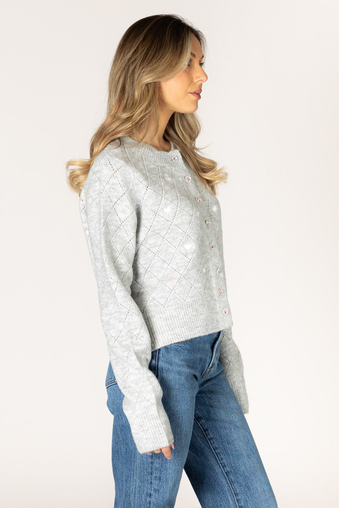 A woman in profile wearing a light gray textured knit cardigan sweater and blue jeans, standing against a plain off-white studio background.