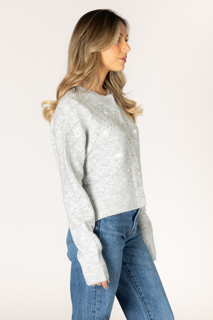 A woman in profile wearing a light gray textured knit cardigan sweater and blue jeans, standing against a plain off-white studio background.