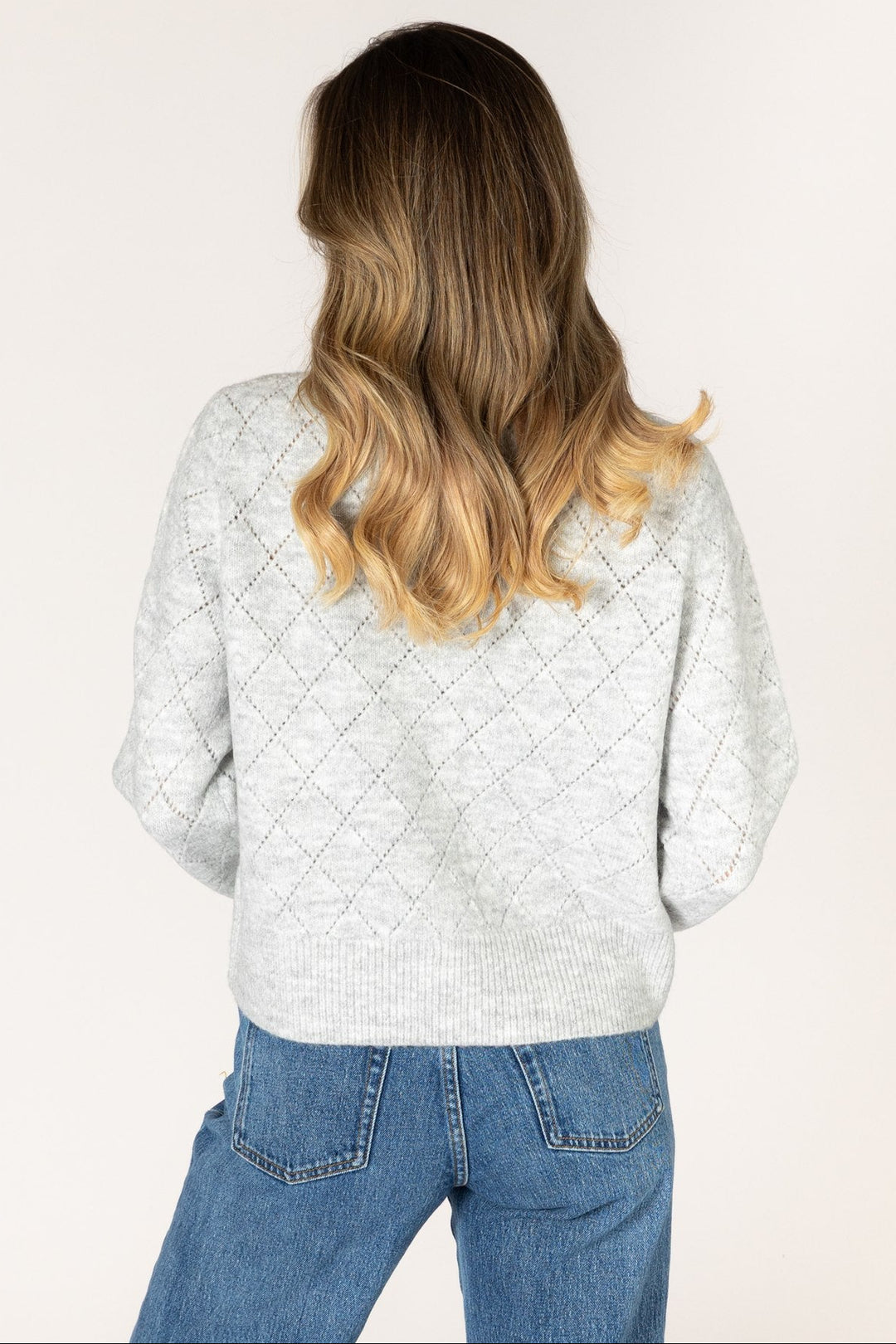 A woman with long blonde-highlighted hair is shown from behind wearing a light gray textured knit sweater and blue jeans against a plain off-white studio background.