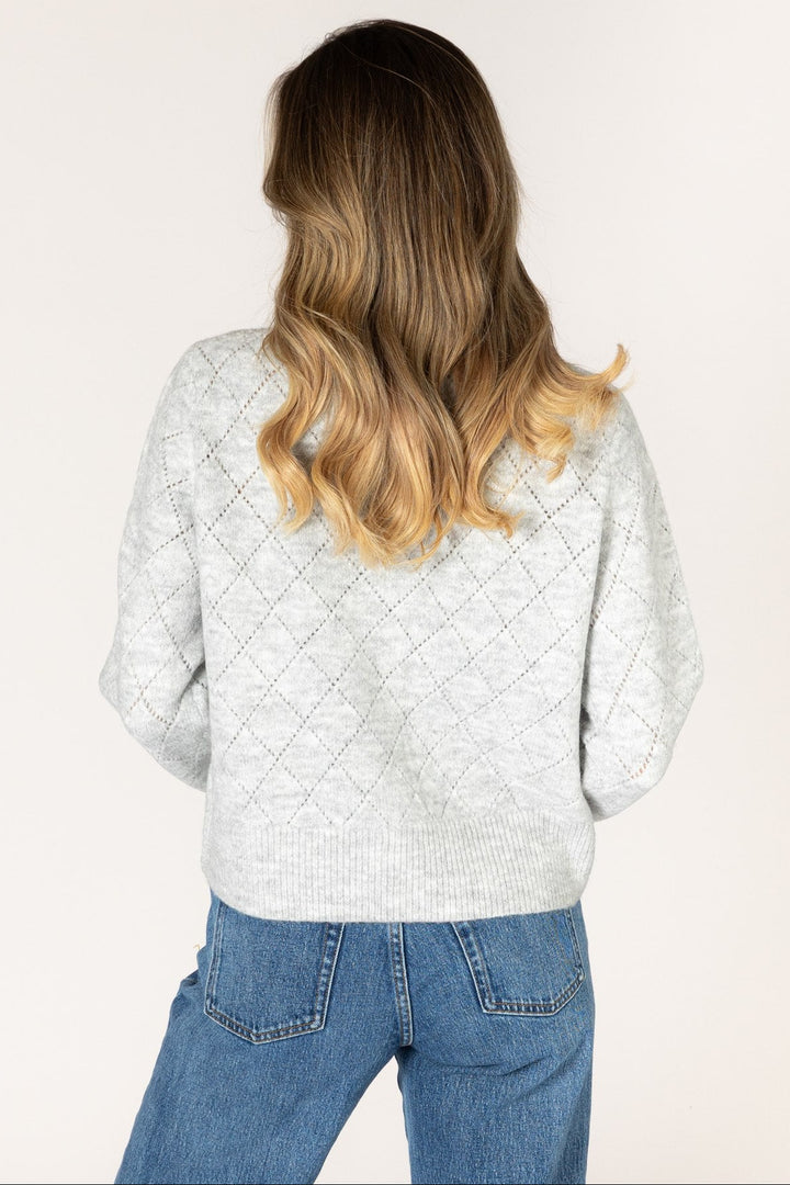 A woman with long blonde-highlighted hair is shown from behind wearing a light gray textured knit sweater and blue jeans against a plain off-white studio background.
