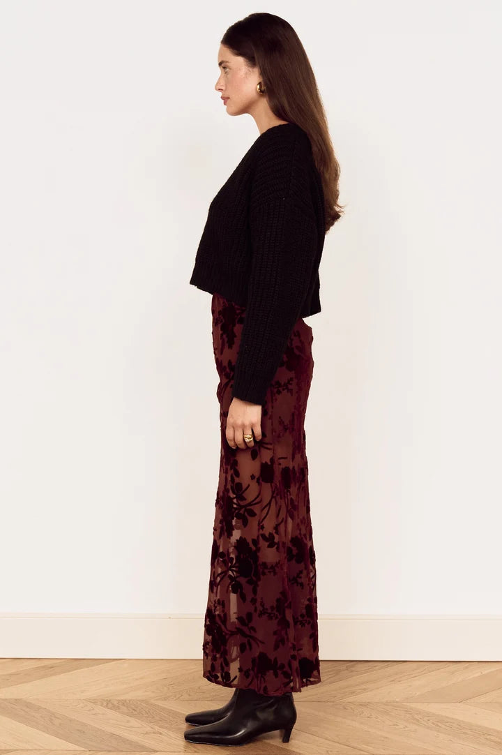 A woman stands in profile wearing a black ribbed cropped sweater and a long burgundy floral sheer skirt with black ankle boots, against a plain white wall and light wood floor.