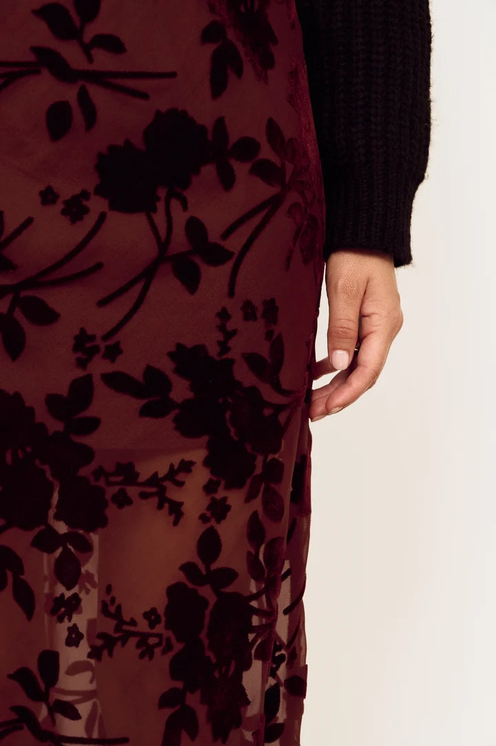 Close-up of a sheer burgundy midi skirt with black floral applique, worn over a black ribbed sweater, shown on a model against a plain off-white background.