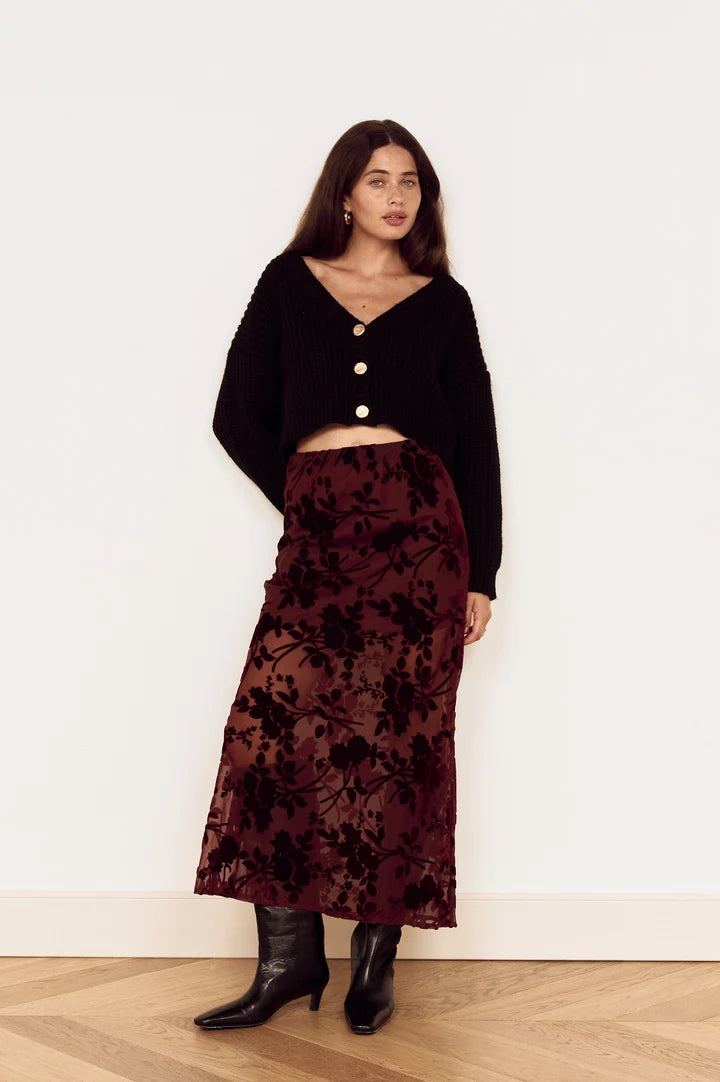 A woman stands in a minimalist room with a white wall and light wood herringbone floor, wearing a black cropped cardigan with gold buttons, a dark red floral velvet midi skirt, and black ankle boots.