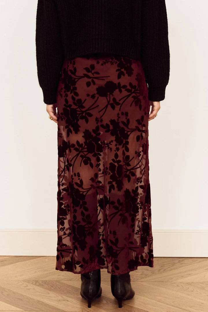 A person shown from the back wearing a long sheer burgundy skirt with a dark floral pattern, paired with a black sweater and black ankle boots, standing on a light wood floor against a white wall.