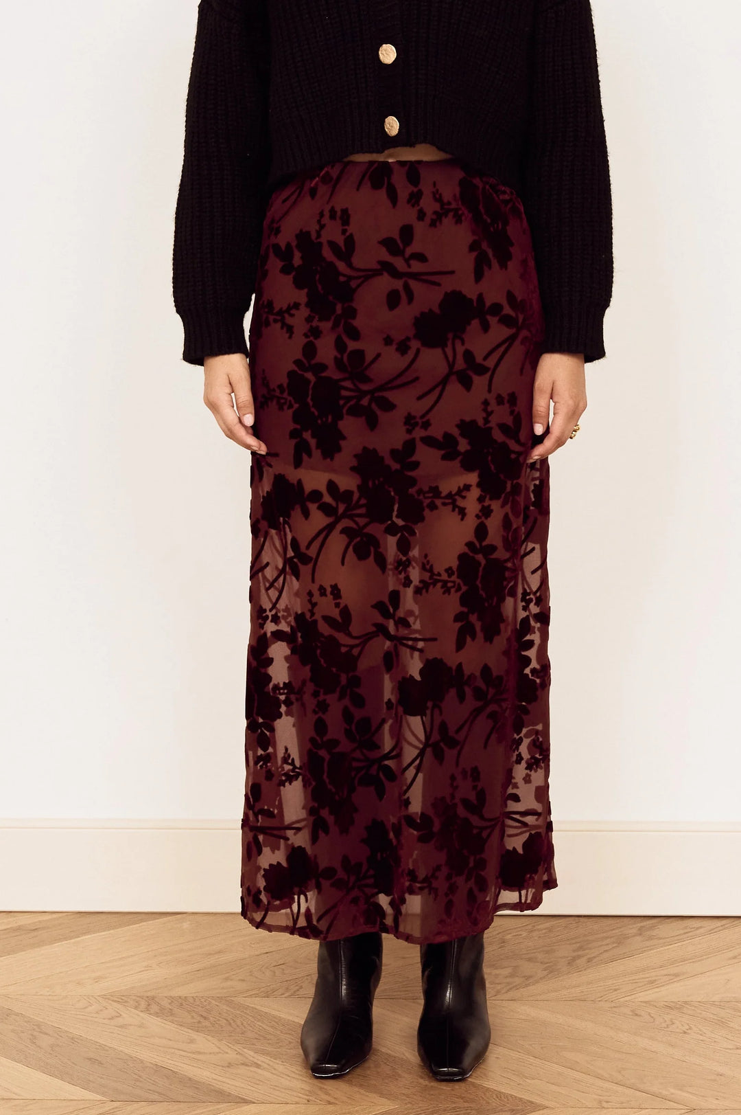 A woman stands against a plain white wall wearing a black long-sleeve button-front top and a dark burgundy floral lace maxi skirt, paired with black ankle boots, on a light wood herringbone floor.