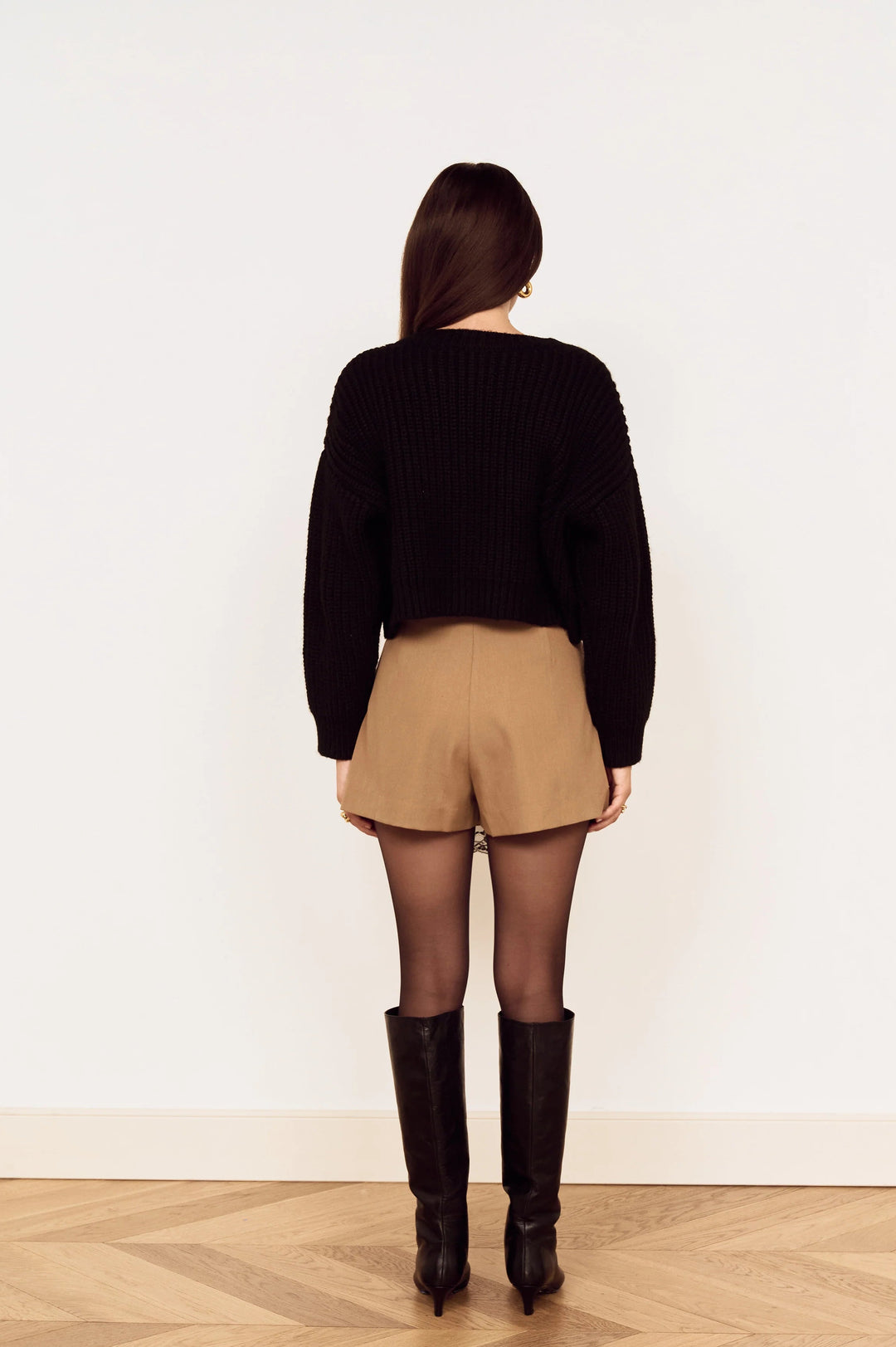 A woman viewed from behind wearing a black ribbed knit sweater, tan shorts, sheer black tights, and black knee-high boots, standing on a light wood herringbone floor against a plain white wall.