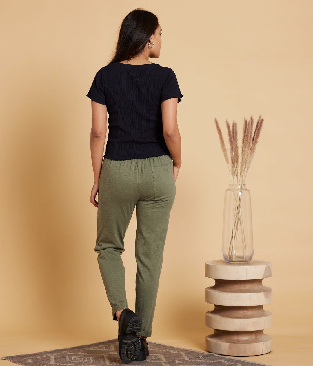 A woman is shown from behind wearing a black short-sleeve top and olive green drawstring pants, standing on a patterned rug next to a decorative wooden pedestal with a glass vase of dried grass against a solid beige background.