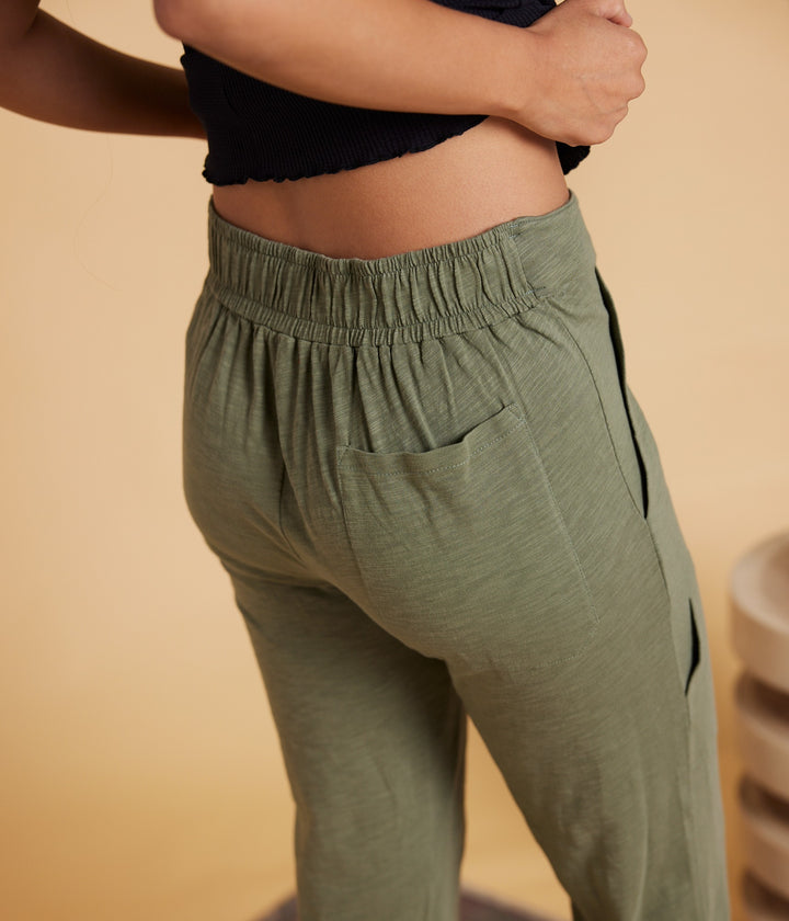 A close-up lifestyle shot of a person wearing olive green athletic jogger pants with an elastic waistband, side pockets, and black side stripes, paired with a black cropped top against a warm beige background.