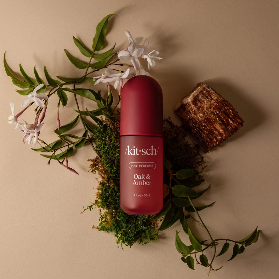 A bottle of Kit Schö Oak &amp; Amber body perfume in a deep red bottle, styled in a flat-lay with green leaves, white blossoms, moss, and a piece of wood on a beige background.