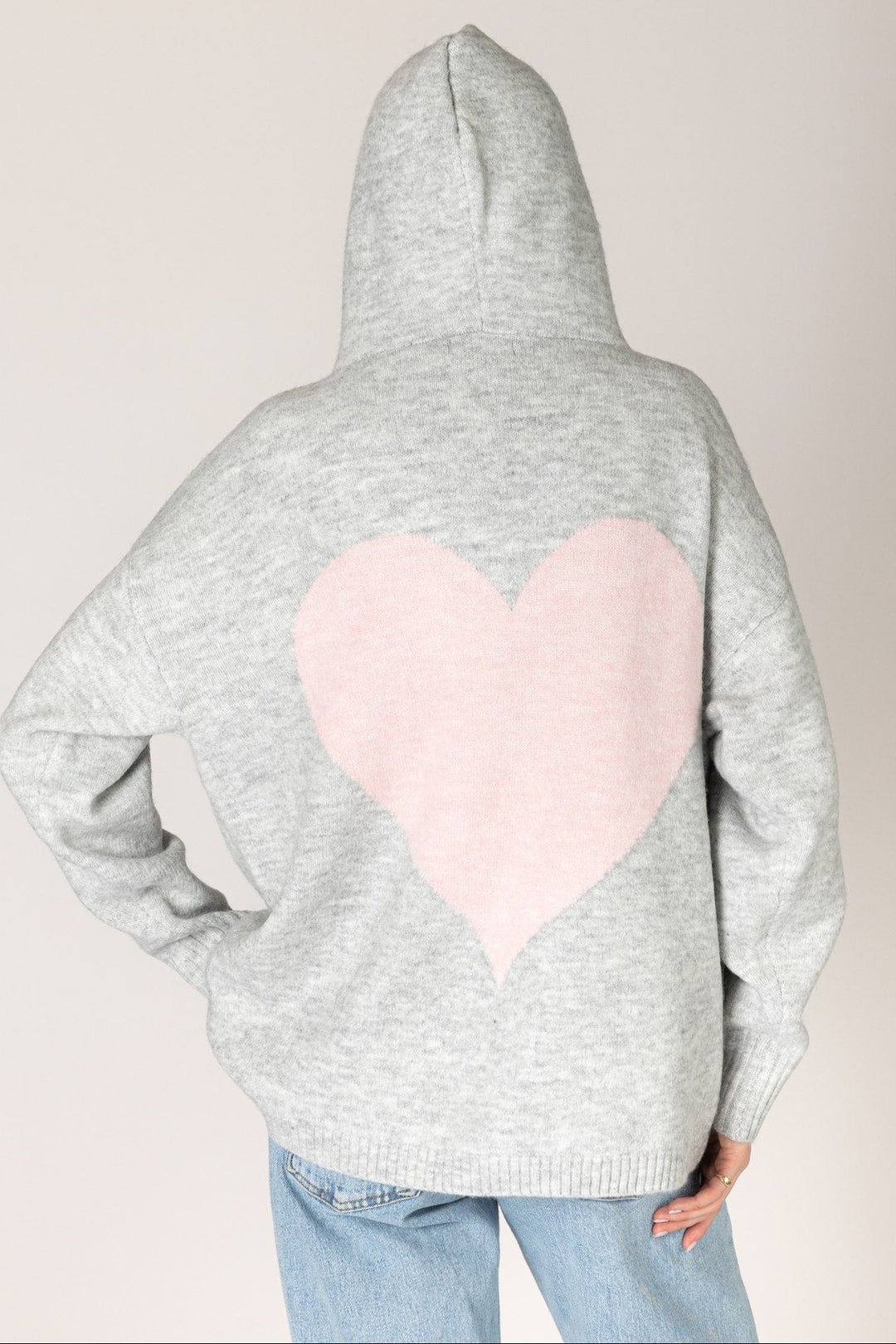 A person shown from the back wearing a light gray heathered hoodie with a large light pink heart patch on the back, paired with light blue jeans, against a plain light beige background.