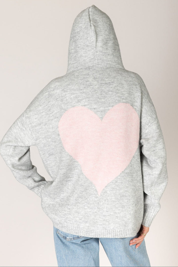 A person shown from the back wearing a light gray heathered hoodie with a large light pink heart patch on the back, paired with light blue jeans, against a plain light beige background.