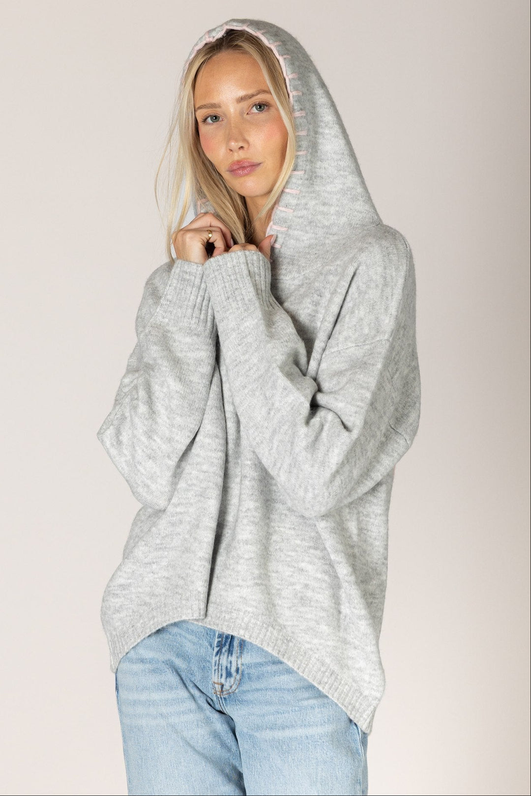 A woman wearing a light gray heathered oversized knit hoodie sweater and light blue jeans, posing against a solid light beige studio background.