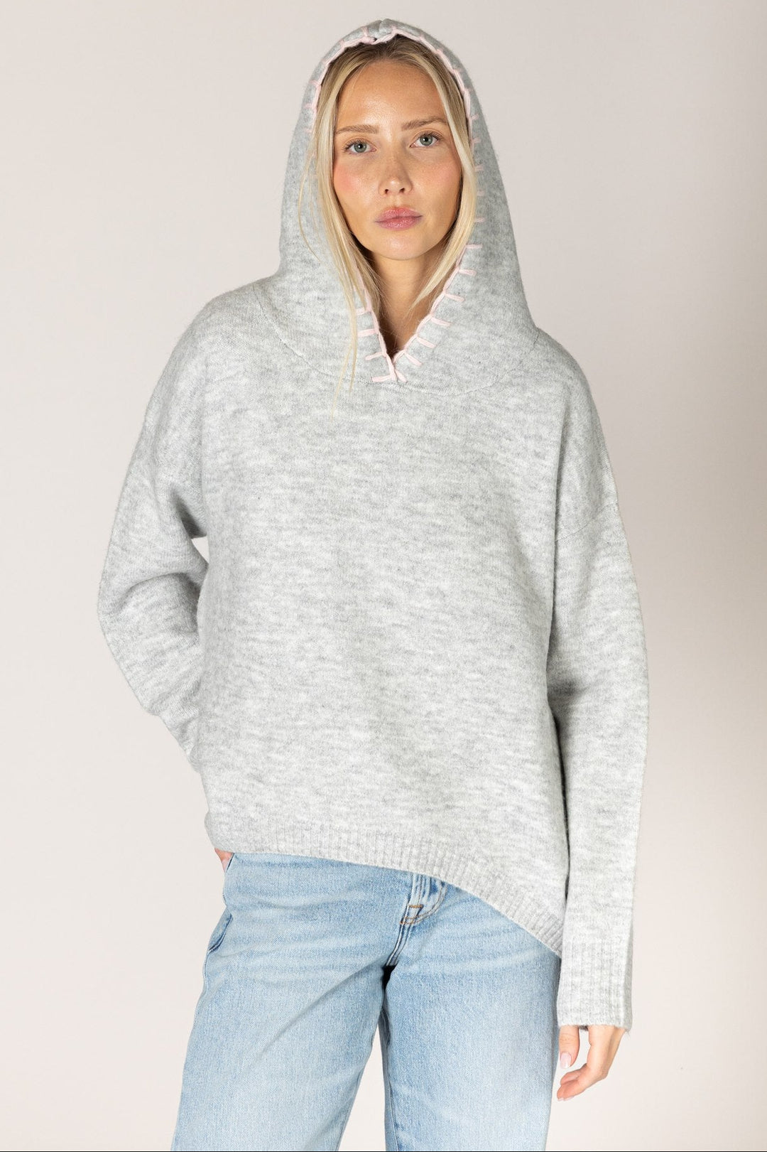 A woman wearing a light gray heathered knit pullover hoodie with a wide collar and drawstring hood, paired with light blue jeans, against a plain light gray studio background.