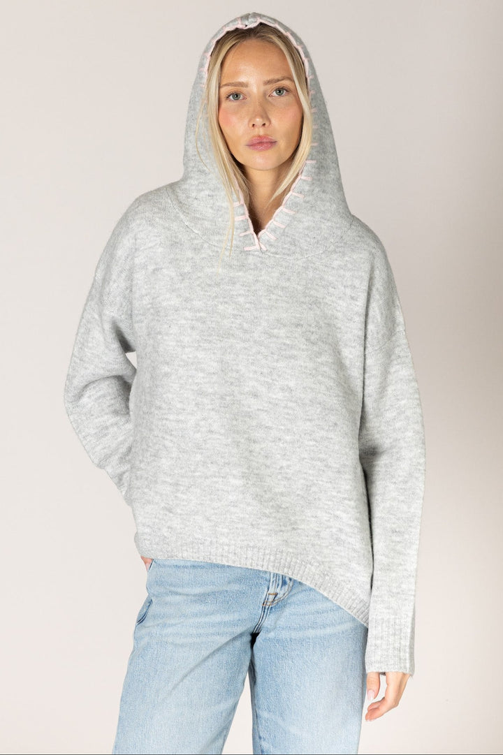 A woman wearing a light gray heathered knit pullover hoodie with a wide collar and drawstring hood, paired with light blue jeans, against a plain light gray studio background.
