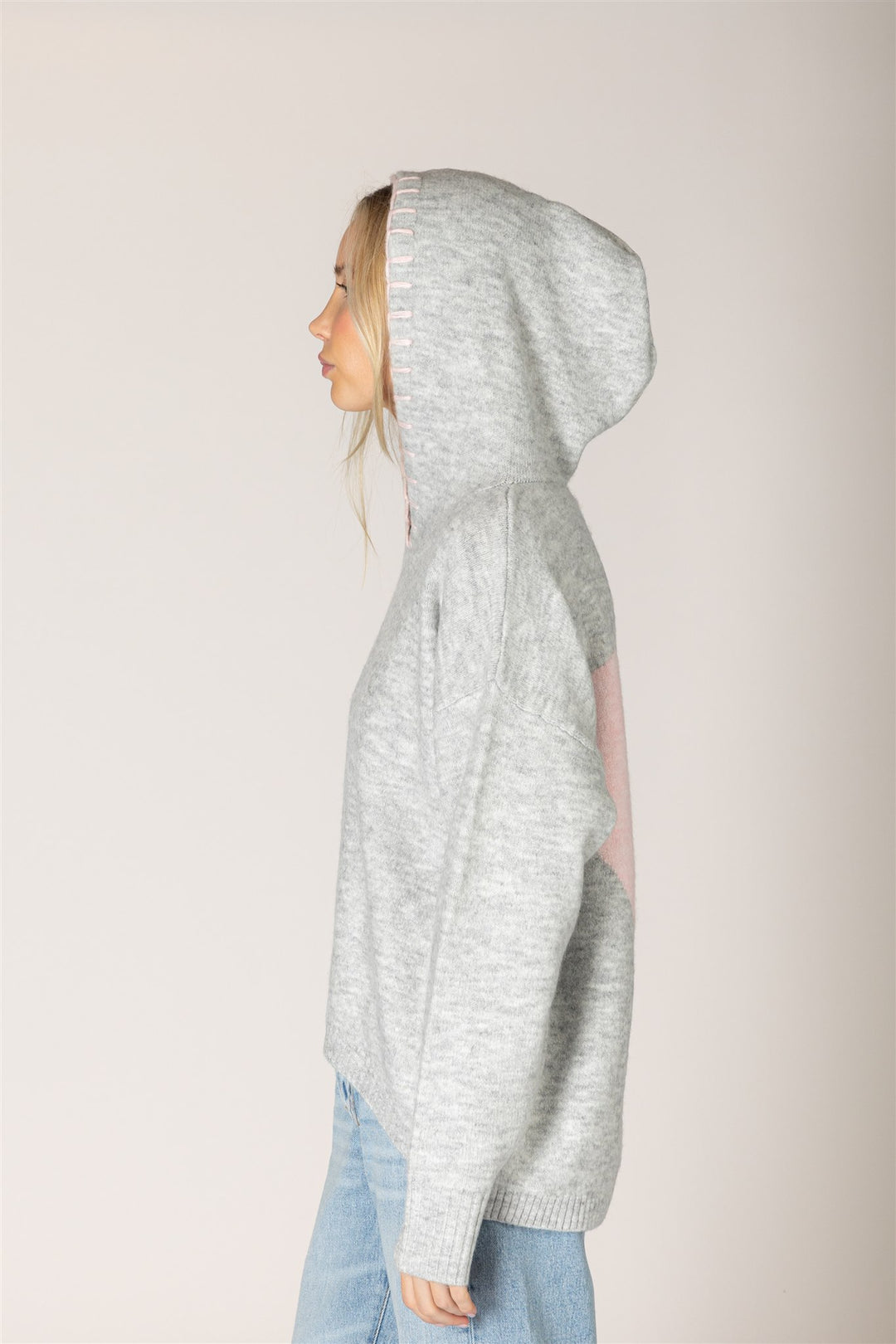 A woman shown in profile wearing a light heather gray knit hoodie with a pink patch on the back, paired with light blue jeans, against a solid light beige background.
