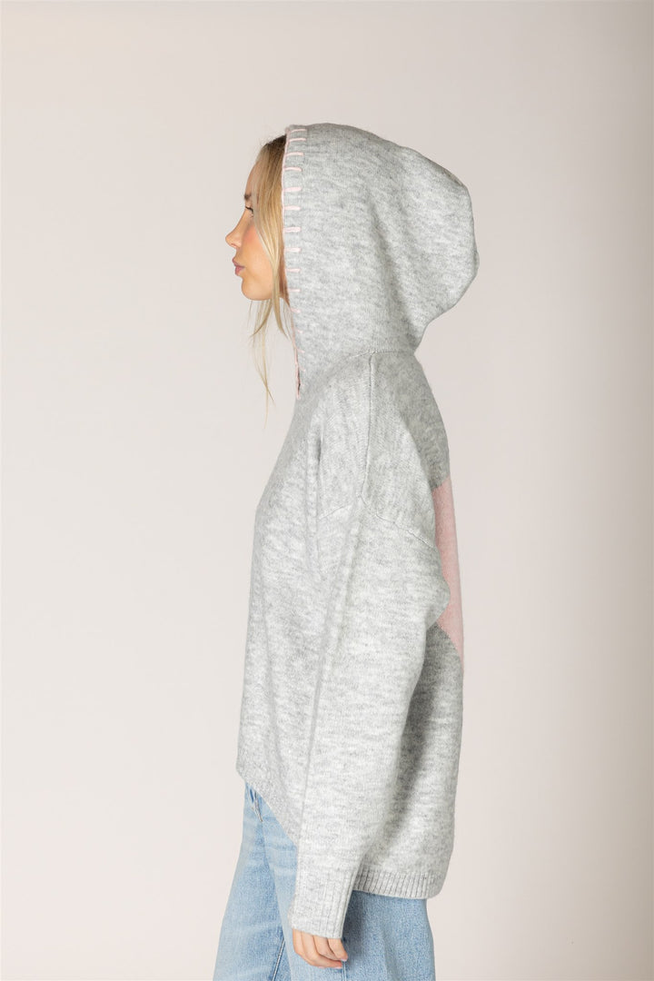 A woman shown in profile wearing a light heather gray knit hoodie with a pink patch on the back, paired with light blue jeans, against a solid light beige background.