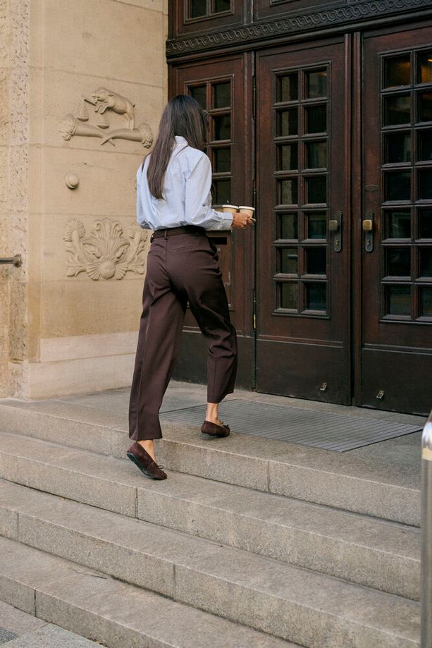 A woman seen from behind walking down stone steps toward dark wooden double doors, wearing a light blue button-up shirt, dark brown tailored trousers, and dark loafers while holding a takeaway coffee cup.