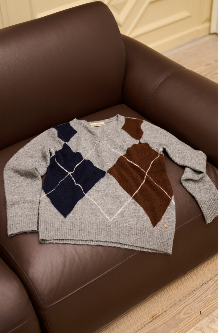 A gray knit sweater with large argyle diamond panels in navy blue, brown, and cream, laid flat on a brown leather sofa.