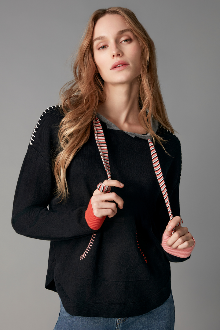 A woman modeling a black long-sleeve knit top with pink and white striped drawstrings and white embroidered details on the shoulders, paired with blue jeans against a solid gray studio background.