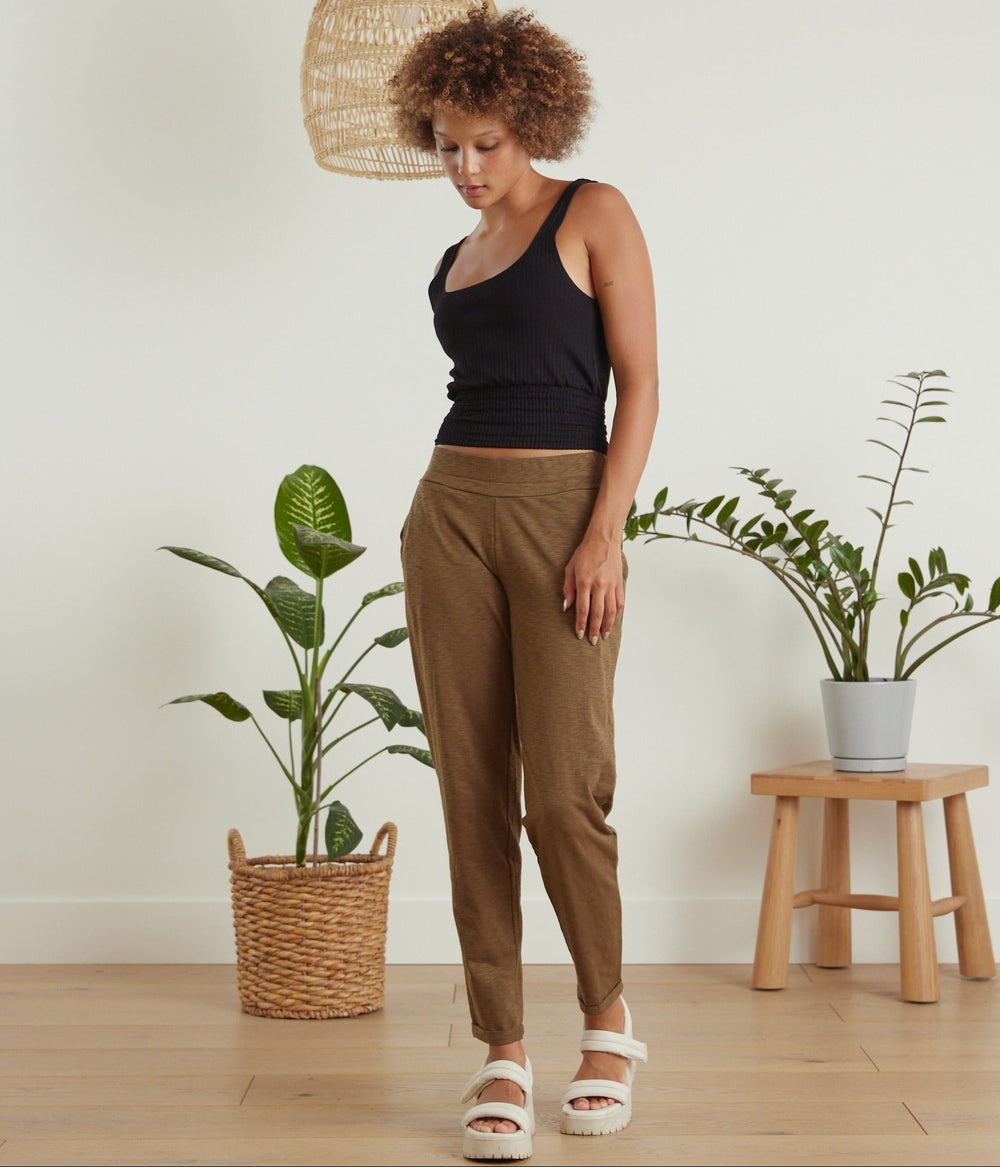 A woman stands in a bright room wearing brown tapered pants, a black tank top, and white slide sandals. The scene includes potted plants and a small wooden stool in the background.