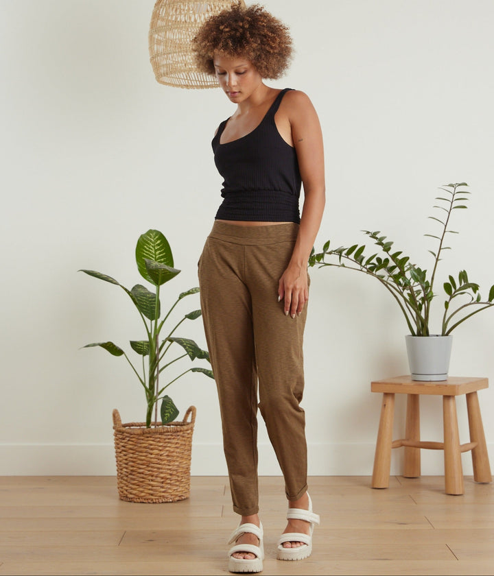 A woman stands in a bright room wearing brown tapered pants, a black tank top, and white slide sandals. The scene includes potted plants and a small wooden stool in the background.