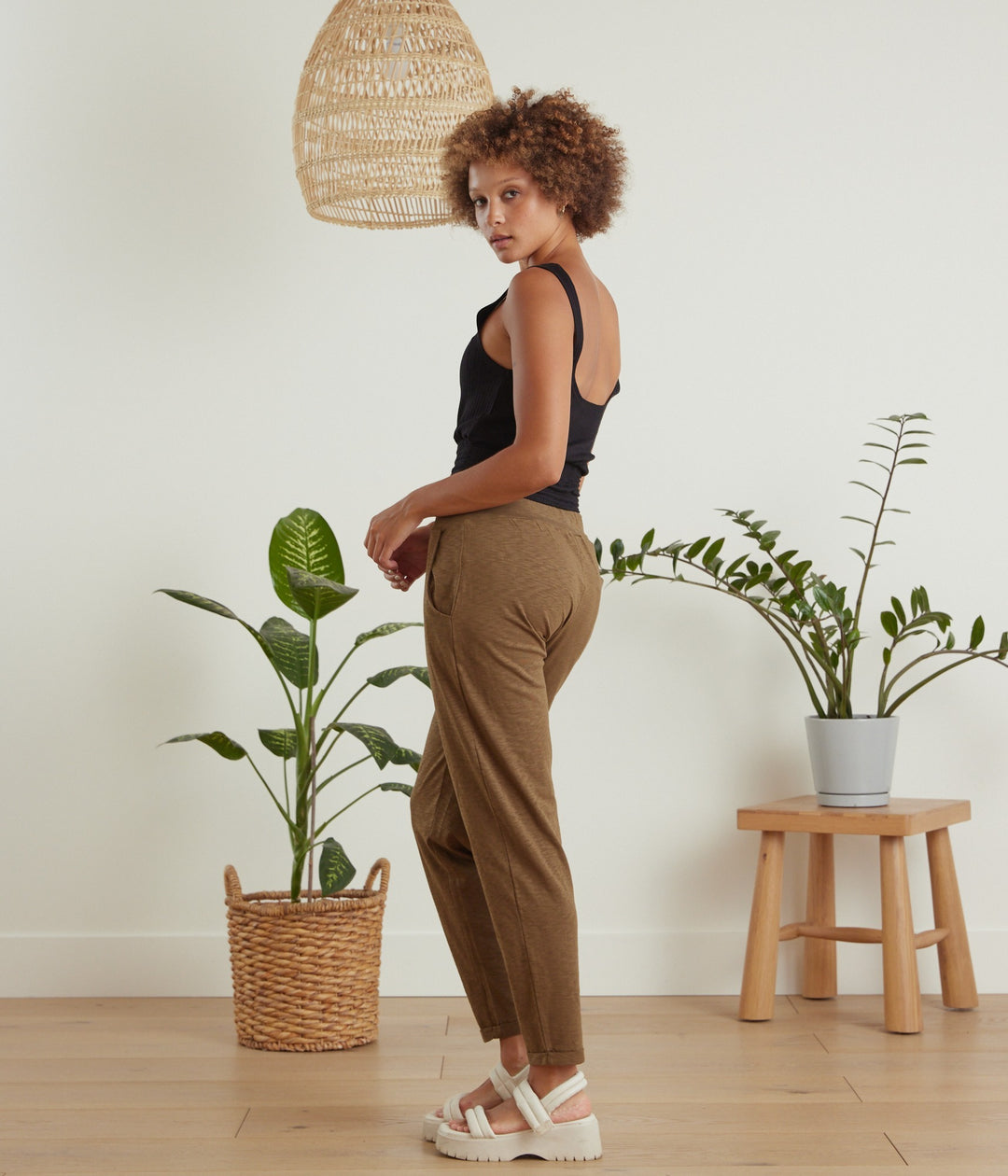 A woman stands in profile wearing a black tank top and brown wide-leg cropped pants, posed in a bright room with potted plants, a wooden stool, and a hanging woven pendant light.