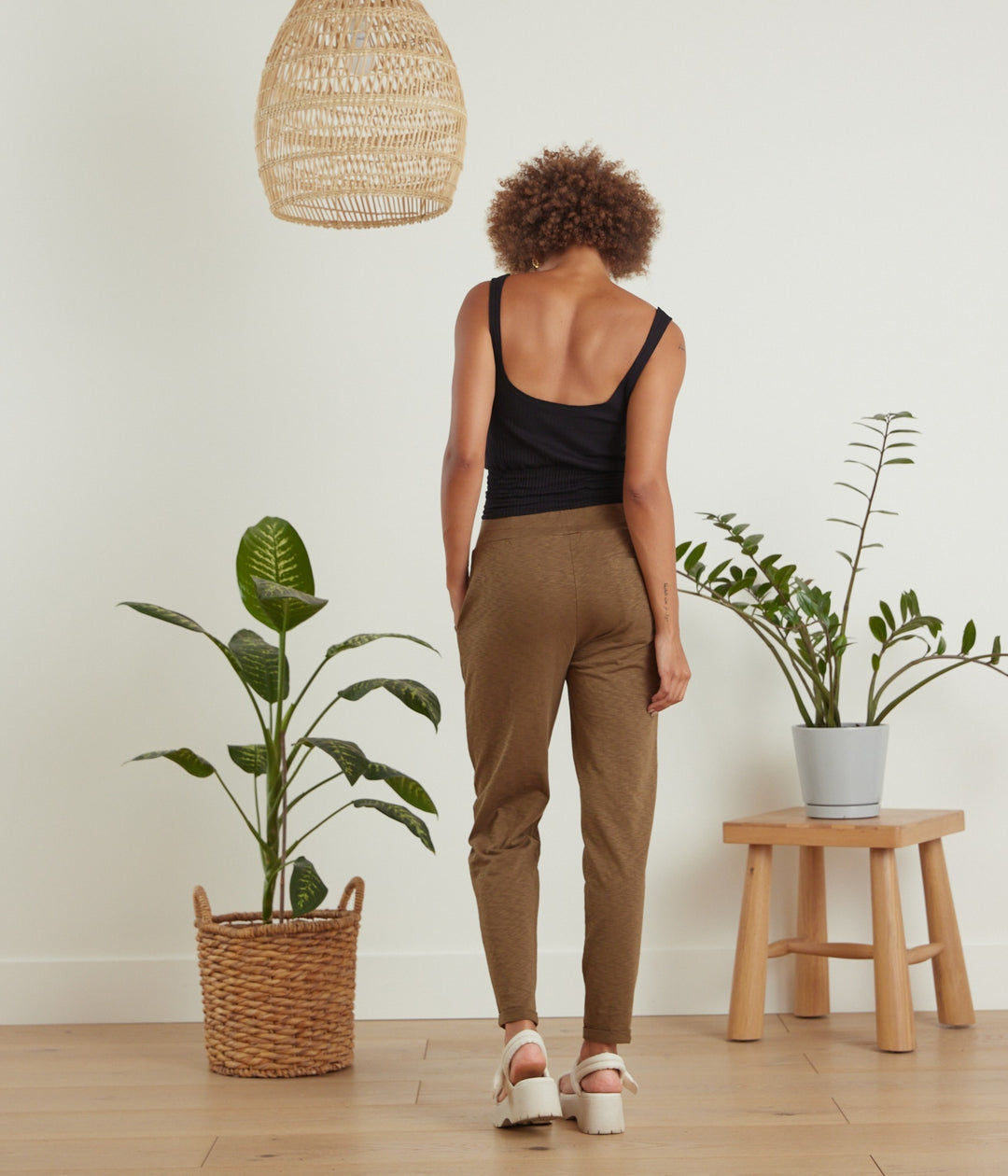 A woman stands with her back to the camera wearing brown tapered trousers and a black tank top, in a bright room with potted plants, a wooden stool, and a hanging wicker lamp.