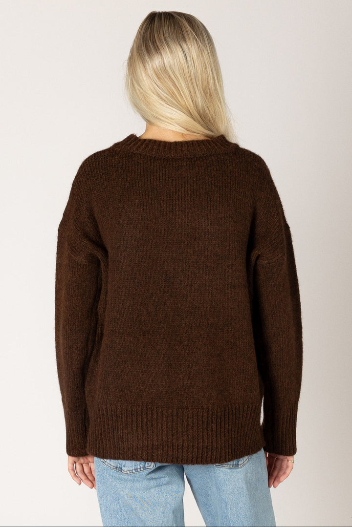 A woman viewed from behind wearing a dark brown knit sweater and light blue jeans against a plain light gray studio background.