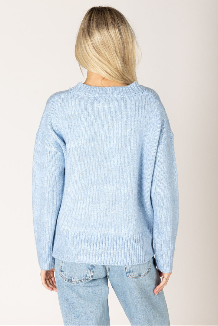 A woman viewed from the back wearing a light blue knit sweater and light blue jeans against a plain light beige studio background.