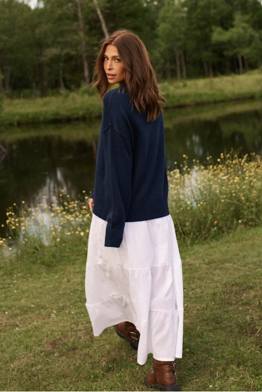 A woman stands outdoors near a pond and trees, wearing a navy blue long-sleeve top and a long white skirt, with brown boots. The background shows grass, yellow wildflowers, water, and dense green trees under a cloudy sky.