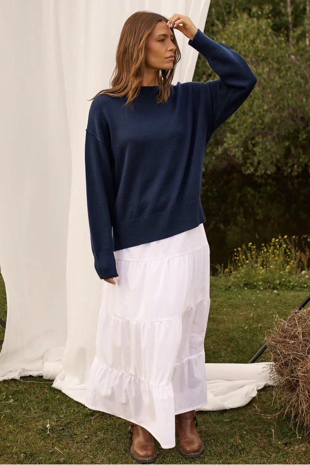 A woman stands outdoors wearing a navy blue long-sleeve crewneck sweater and a long white tiered skirt, with brown boots. A large white draped fabric backdrop is behind her, with grass and trees in the background.