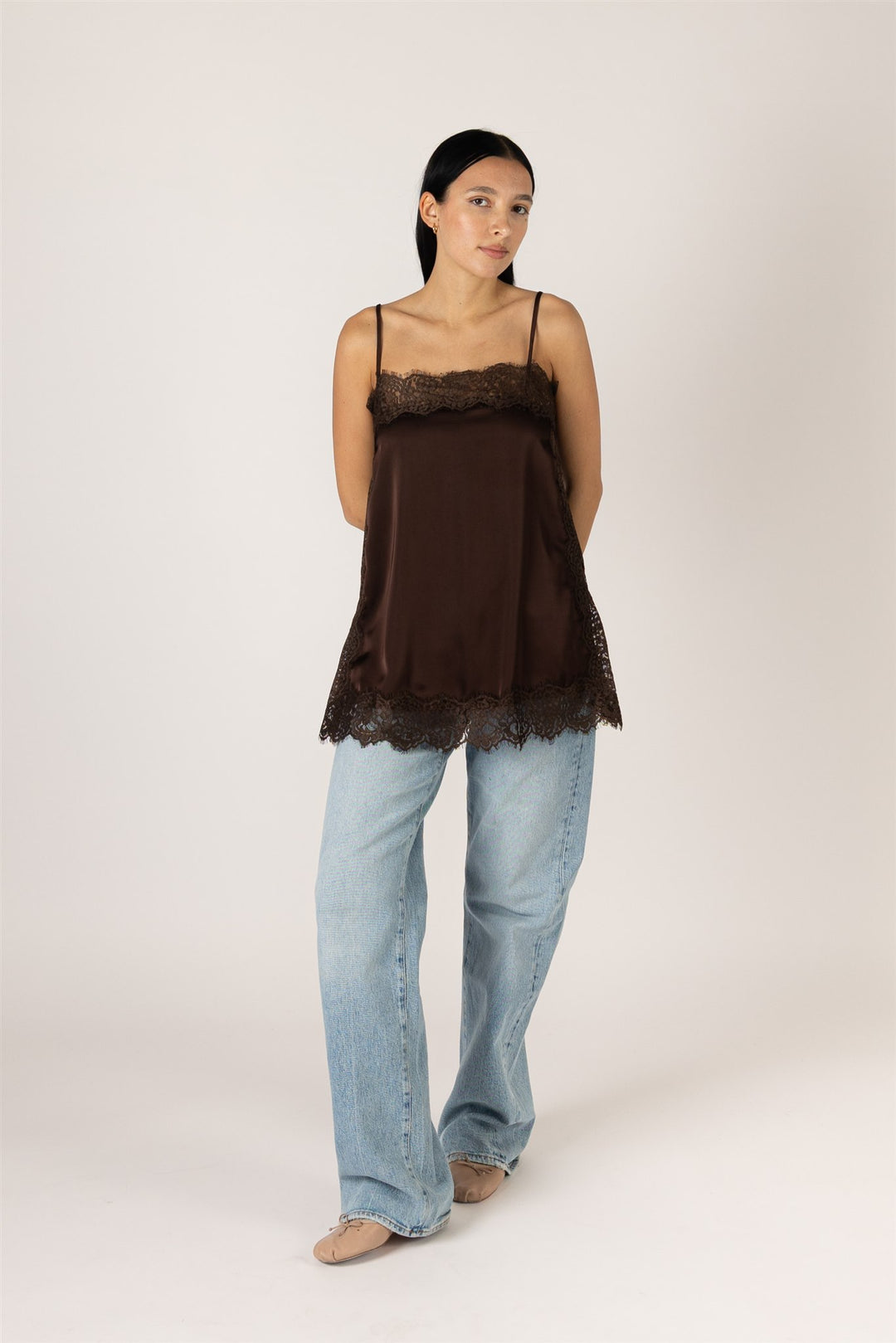 A woman modeling a dark brown camisole top with lace trim, paired with light blue jeans, against a plain light beige studio background.