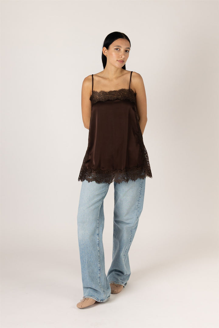 A woman modeling a dark brown camisole top with lace trim, paired with light blue jeans, against a plain light beige studio background.