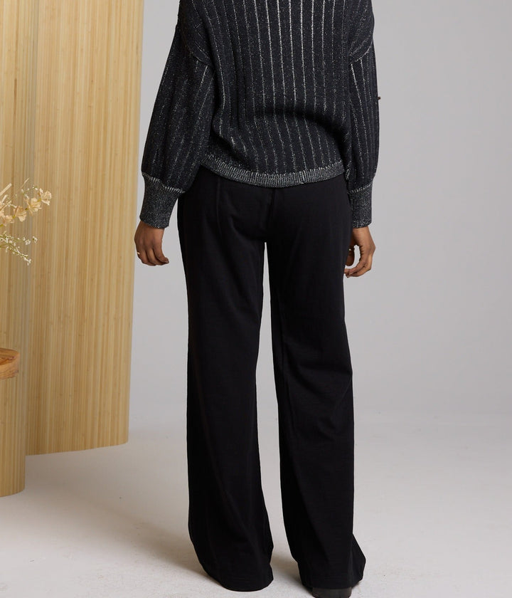 Person wearing a black pinstripe jacket and black pants in a minimalistic setting with a wooden stool and vase.