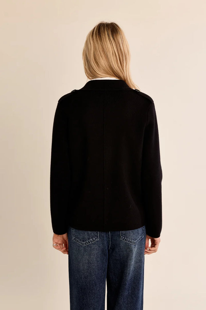 A woman viewed from behind wearing a black long-sleeve knit jacket with shoulder epaulets and blue jeans, set against a plain beige studio background.