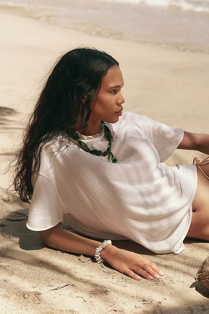 A woman lying on a sandy beach wearing an oversized white short-sleeve top, a green beaded necklace, and a white beaded bracelet.