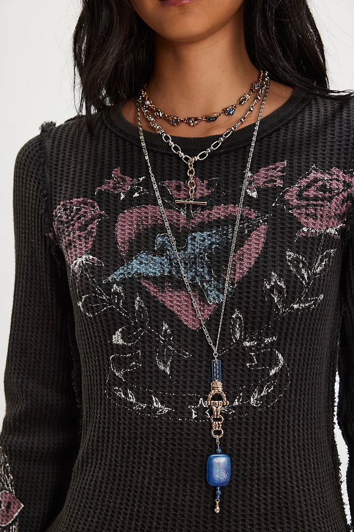 A close-up of a person wearing a black textured knit top with a pink and blue rose graphic, layered with multiple silver and gold necklaces, including a long pendant necklace with a blue bead.