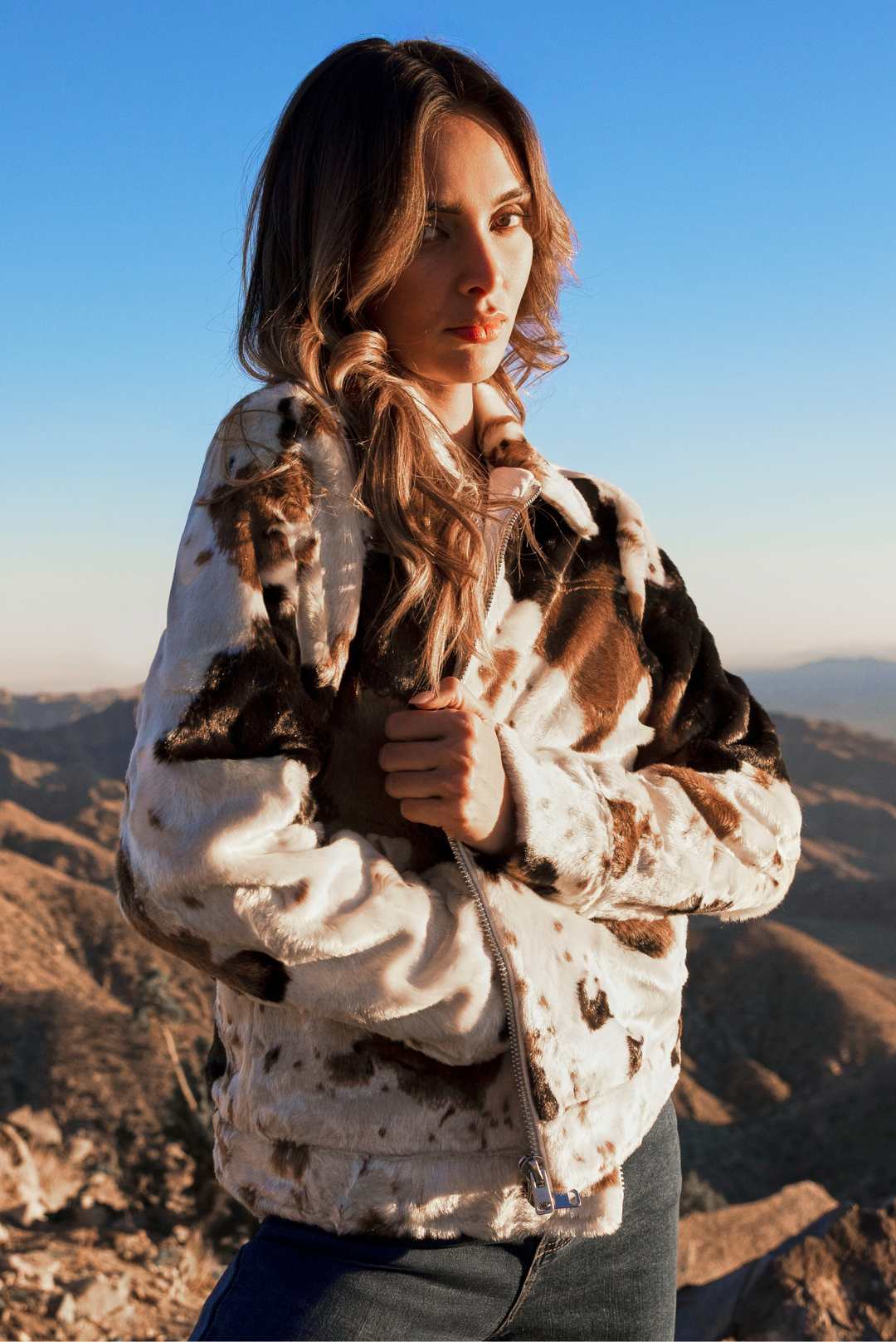 A woman wearing a brown and white cow-print faux fur jacket, standing outdoors against a clear blue sky and mountainous landscape.