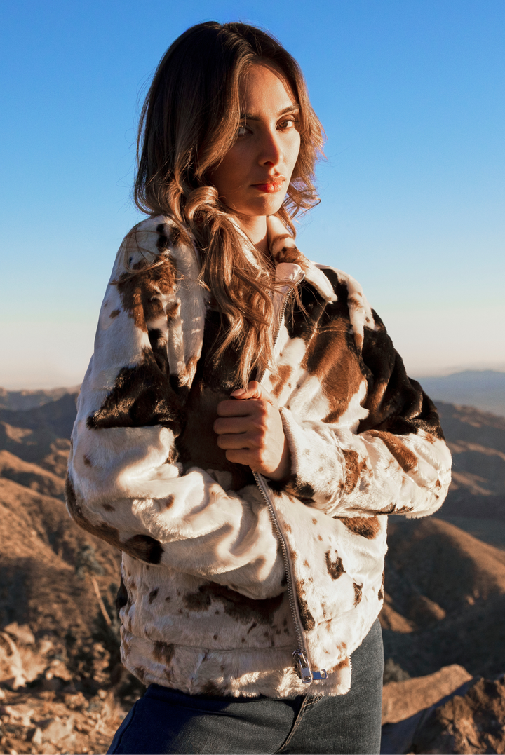 A woman wearing a brown and white cow-print faux fur jacket, standing outdoors against a clear blue sky and mountainous landscape.