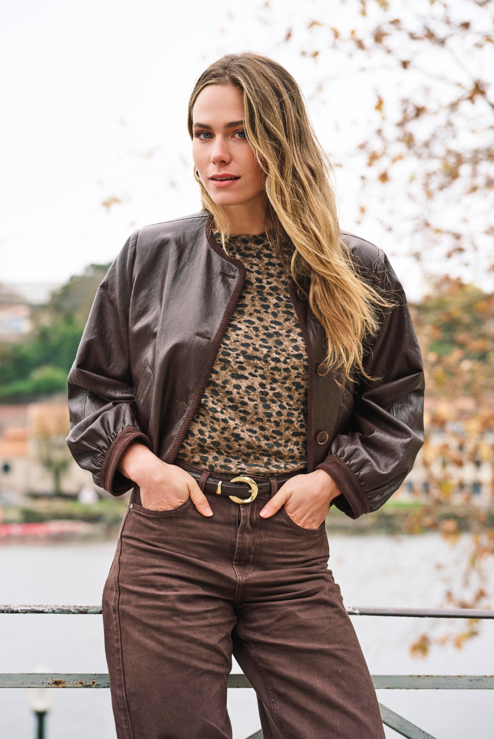 A woman wearing a dark brown faux-leather bomber jacket over a leopard-print top and brown pants, standing outdoors near a railing with a blurred background of water and trees.