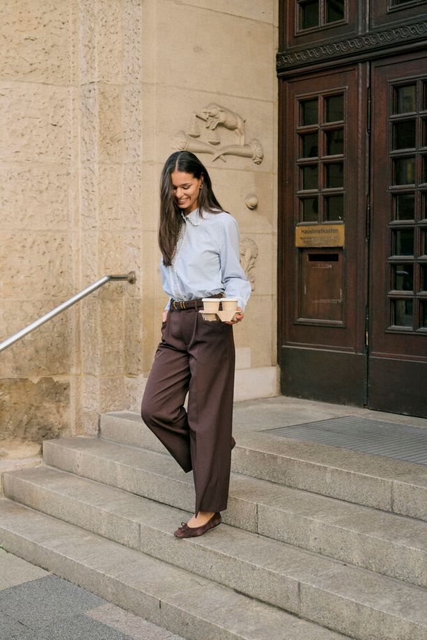 A woman in a light blue button-down shirt and dark brown wide-leg trousers walks down stone steps holding a coffee cup, posing in front of a stone building with dark wooden doors.