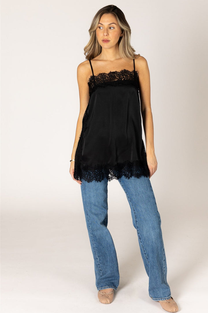 A woman modeling a black camisole top with lace trim at the neckline and hem, paired with blue jeans and beige shoes, against a plain light gray studio background.