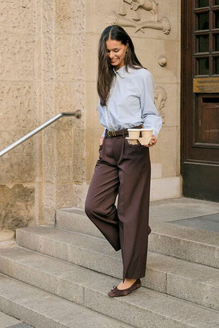 A woman in a light blue button-down shirt and dark brown wide-leg trousers walks down stone steps holding a coffee cup, posing in front of a stone building with dark wooden doors.